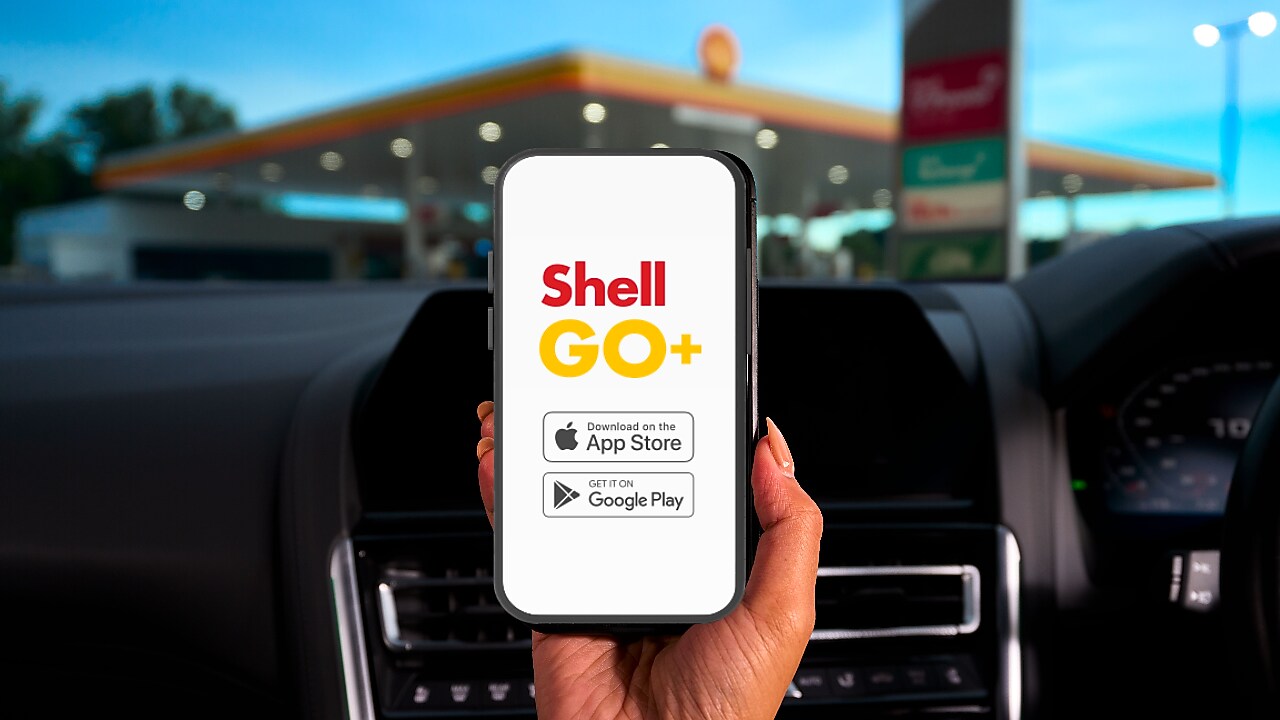 Persona sostiene un teléfono móvil con la app Shell GO+ abierta, mostrando las opciones para descargar desde App Store y Google Play, dentro de un auto frente a una estación de servicio Shell. Promoción digital de fidelidad y recompensas de Shell.