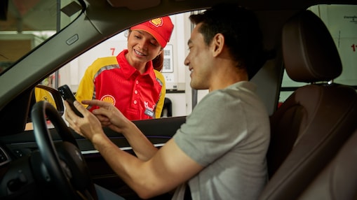 Cliente sentado dentro de su automóvil interactúa con un trabajador de Shell a través de la ventana del conductor, mostrando su teléfono móvil. Programa de fidelización y pago móvil en gasolineras Shell.