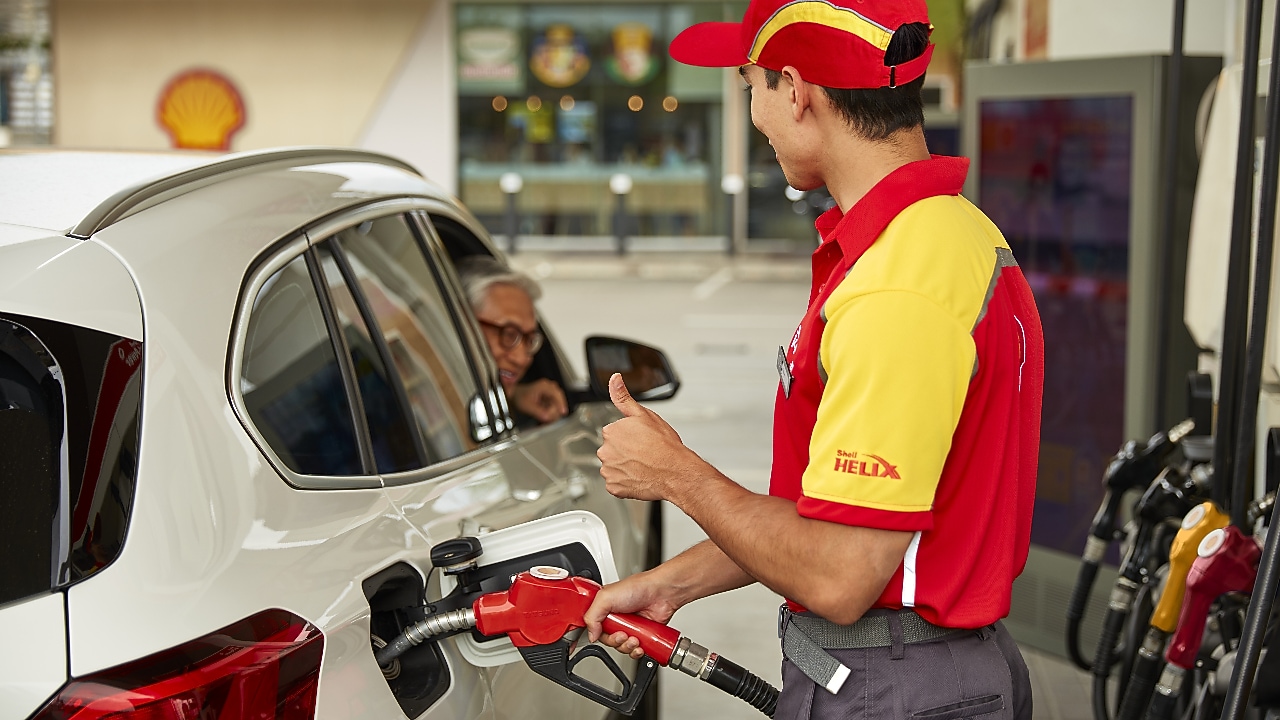 Empleado de Shell con uniforme rojo y amarillo llena el tanque de un vehículo blanco mientras conversa con un cliente en una estación de servicio. Servicio al cliente y abastecimiento de combustible en estaciones Shell.