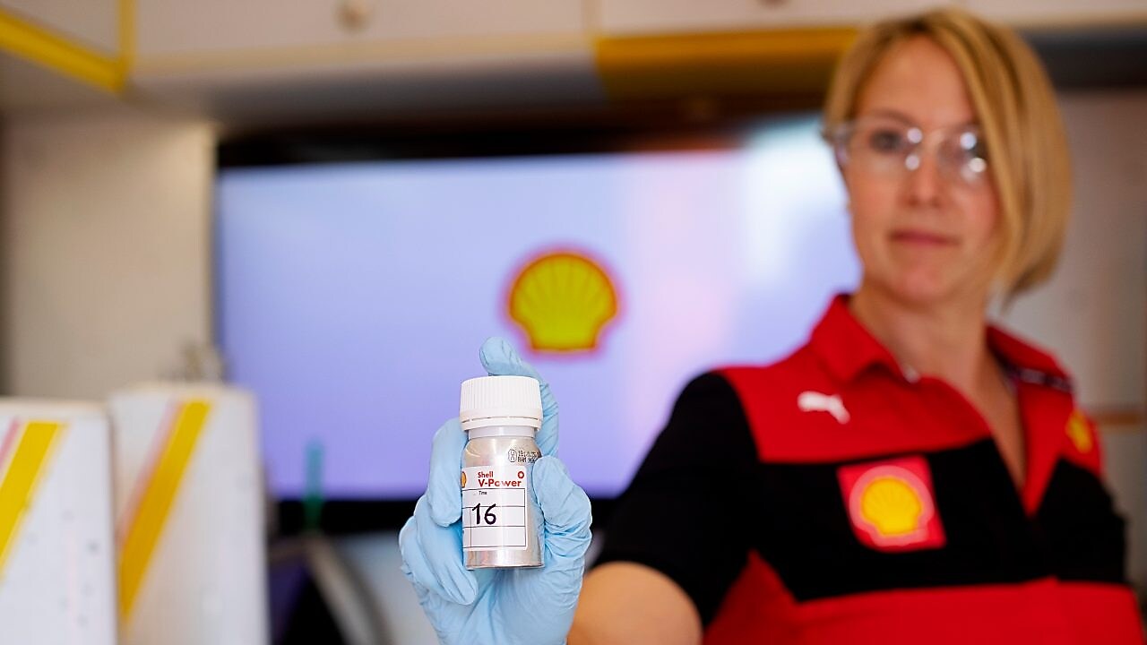 Al fondo, una mujer blanca con cabello rubio sostiene una botella escrita "Shell V-Power"