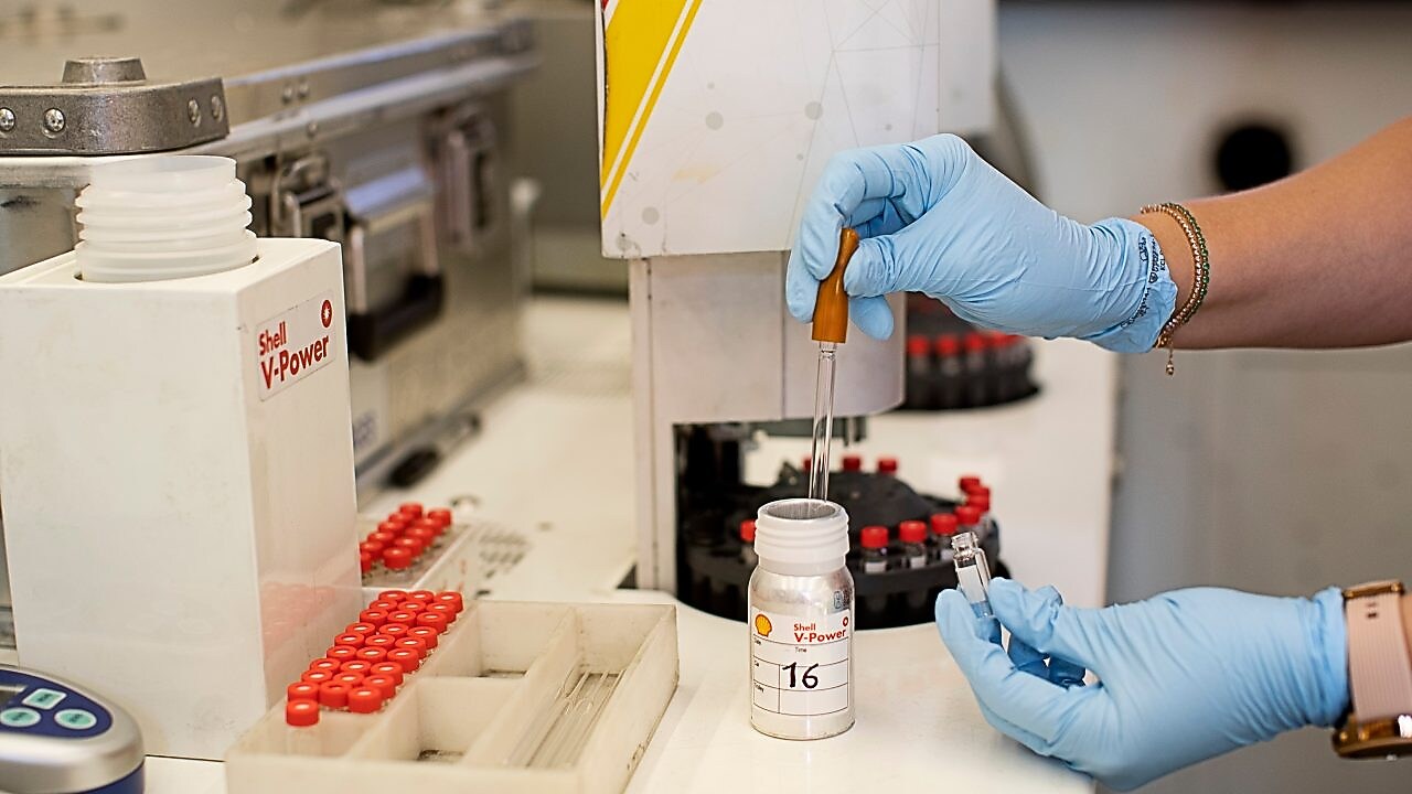 Dos manos con guantes de laboratorio trabajan en una botella escrita "Shell V-Power"