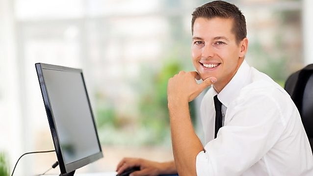 Hombre de pelo castaño sonriendo a la foto en cuanto trabaja en su computadora